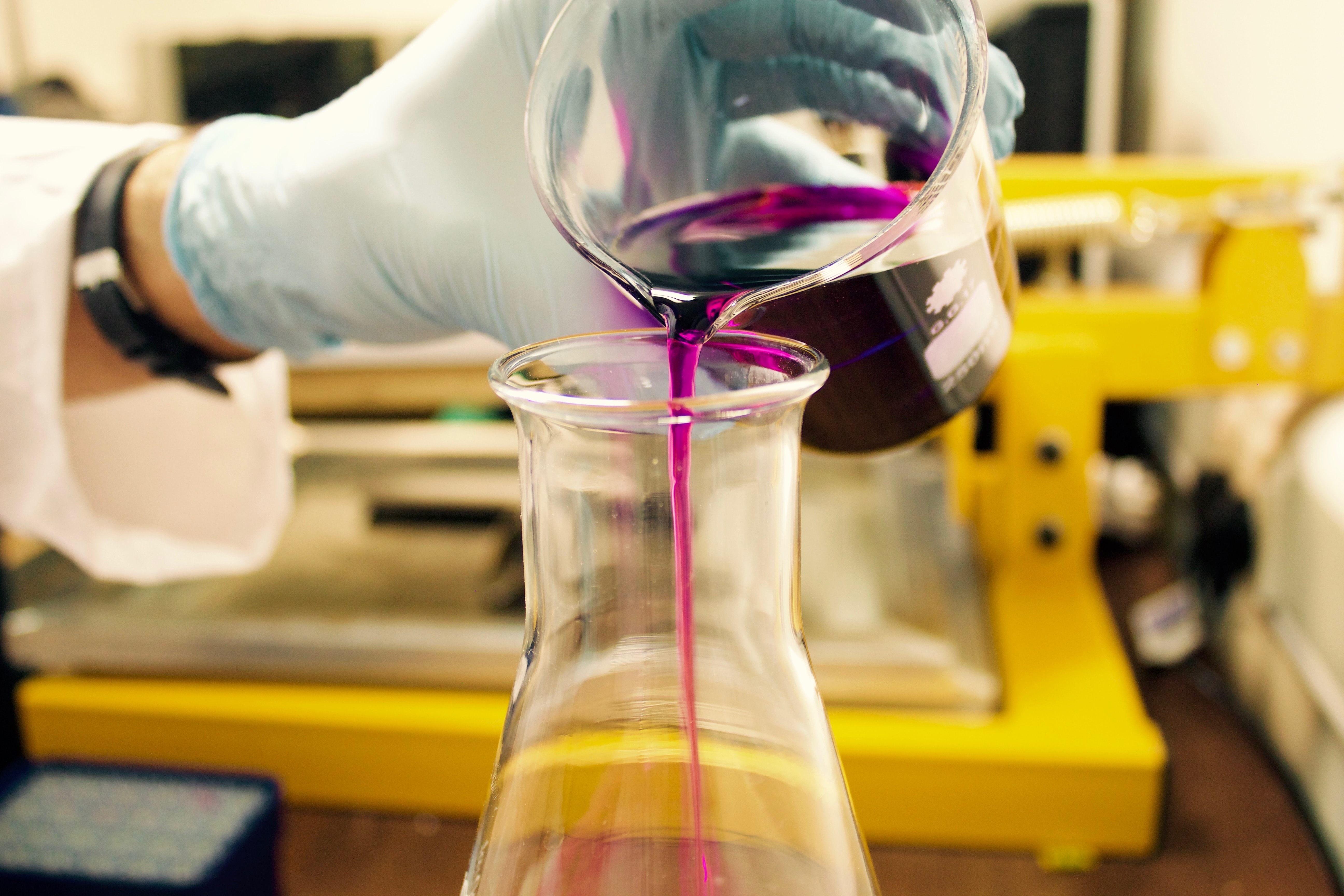 A gloved scientist pours purple liquid from a beaker into a conical flask.