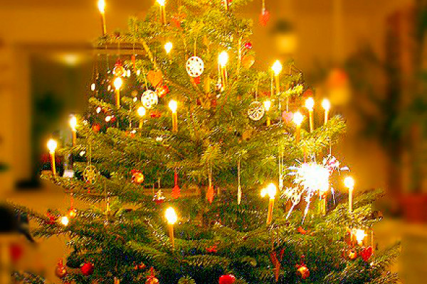 A Danish Christmas tree illuminated with burning candles, adorned with homemade Christmas decorations such as red hearts, white paper snowflakes, a golden star at the top, and gifts stacked underneath.
