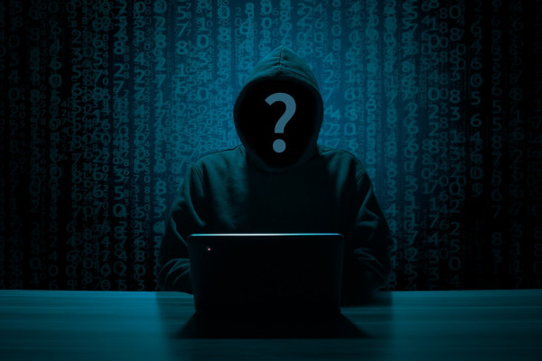 The image shows a hooded person hacking a laptop.
