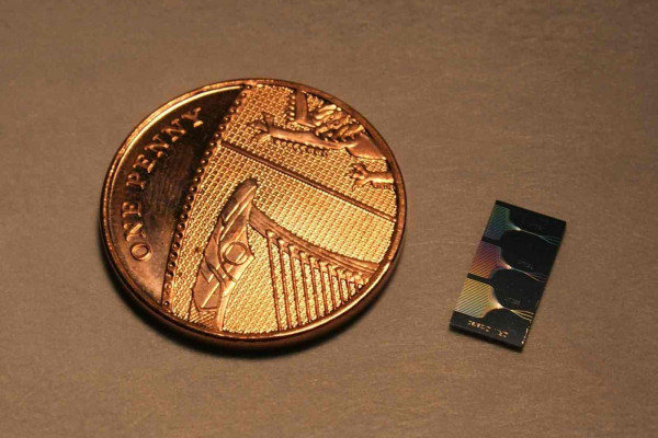 The photonic chip next to a UK penny. The chip contains micrometer and submicrometer features and guide light using a network of waveguides. The output of this network can be seen on the surface of the chip.