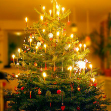 A Danish Christmas tree illuminated with burning candles, adorned with homemade Christmas decorations such as red hearts, white paper snowflakes, a golden star at the top, and gifts stacked underneath.