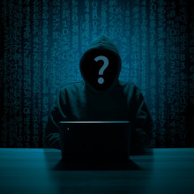 The image shows a hooded person hacking a laptop.